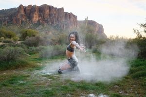 Smiling woman crouches low and reaches toward the camera as powder billows around her in a scenic desert setting.