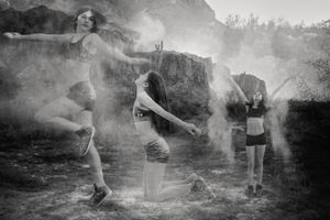 Black and white photo of three dancers posing in dramatic stances amid clouds of powder in front of rugged desert rocks.