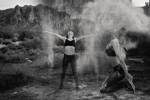 Black and white image of two women with raised arms and powder clouds dancing before towering desert rock formations.