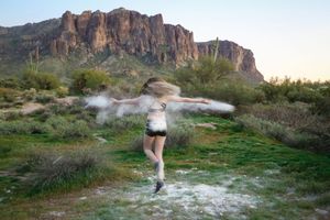Blurred motion of a dancer spinning with trailing powder on a green desert hillside beneath a rocky mountain at dusk.