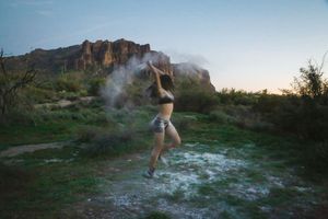 Dancer in athletic wear leaps through a cloud of white powder on a grassy desert trail with rocky mountains behind at dusk.