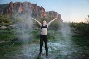 Woman in a black sports bra and leggings stands with arms raised as white powder floats around her in front of red desert cliffs.