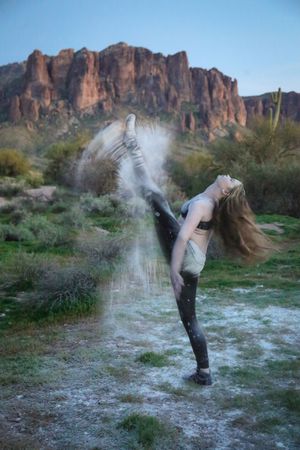 Flexible dancer performs a high standing split kick as white powder bursts in a green desert with red rock formations behind.