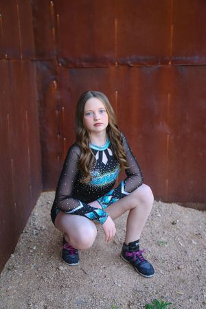 Young cheerleader in a black and teal rhinestone uniform and sneakers crouches against a rusty metal wall on a dirt ground.