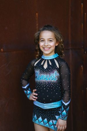 Smiling young cheerleader with curly hair poses in a black and teal Element cheer uniform in front of a rusty metal wall.