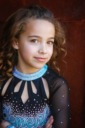 Portrait of a young girl with curly hair and a calm expression in a black and teal Elements cheer uniform by a rusty wall.