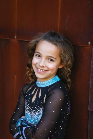 Close-up portrait of a smiling girl with curly brown hair wearing a black and teal sparkly cheer uniform against rusty metal.