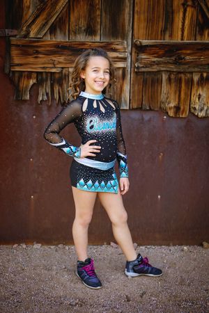 Smiling young cheerleader in a black and teal sparkle uniform and sneakers poses with hand on hip in front of a rustic wooden barn door.