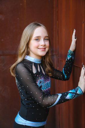 Young cheerleader with long light brown hair smiles while resting her hands on a rusty wall, wearing a black and teal sparkle uniform.