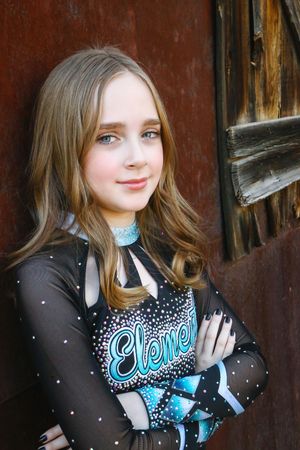 Portrait of a cheerleader with long wavy hair and crossed arms in a black rhinestone uniform with teal Elements lettering by a rustic wall.