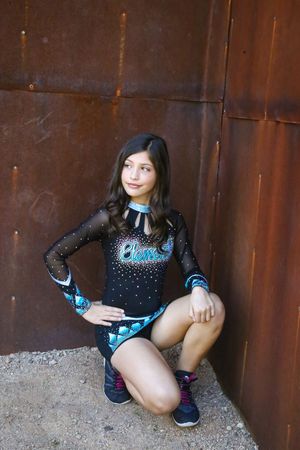 Cheerleader in a black and teal rhinestone uniform crouches on gravel beside a rusty metal wall while glancing to the side.