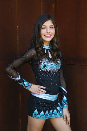 Smiling cheerleader with long dark wavy hair poses with a hand on her hip in a black and teal Elements uniform against a rust-colored wall.