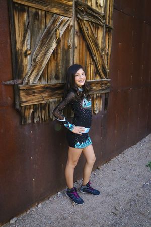 Cheerleader in a black and teal sparkle uniform and sneakers smiles while leaning against a weathered wooden barn door on a gravel path.