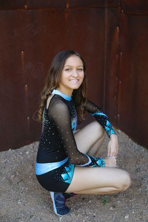Smiling cheerleader in a black and teal sparkly uniform crouching on gravel in front of a rusty metal wall.