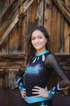 Young cheerleader in a black and turquoise rhinestone uniform posing with hand on hip in front of a rustic wooden barn door.