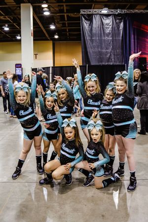 A group of smiling young cheerleaders in black and teal uniforms with matching bows pose together at an indoor competition venue.