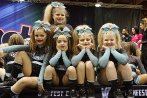 Four young cheerleaders in matching teal and black uniforms with sparkly bows smile and pose together at a cheerleading competition.