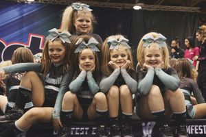 Four young cheerleaders in gray and teal uniforms with big bows pose with hands under their chins at a cheer competition.