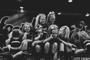 Young cheerleaders in team uniforms and hair bows sit in a row on a stage at a cheer event in a black and white photo.