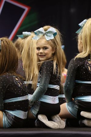 A smiling young cheerleader in a sparkly black and teal uniform with a large glittery bow looks back over her shoulder at a competition.