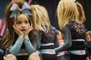 Young cheerleader with a large glitter bow rests her chin on her hands while teammates in sparkly uniforms stand behind her.