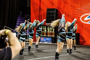 A cheer team in teal and black uniforms performs a synchronized arm-raised routine on an arena floor in front of a red USA banner.