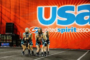 Cheerleaders in teal and black uniforms huddle and lift a teammate during a routine in front of a United Spirit Association banner.