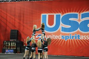 A cheer team performs a stunt on stage in front of a red United Spirit Association banner at a cheerleading competition.
