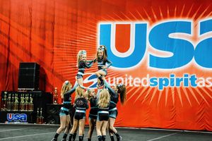 Cheer team performs a stunt as a flyer is lifted high by teammates in front of a red United Spirit Association backdrop.