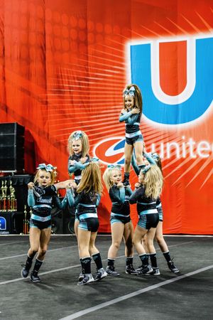 Young cheerleaders with teal bows lift a flyer in a stunt while teammates support below on the competition floor.