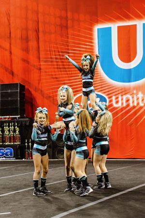 Youth cheer team performs a pyramid stunt as a smiling flyer stands atop teammates in teal and black uniforms.