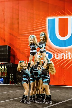 Young cheerleaders in teal and black uniforms hoist two flyers into the air during a competition routine.
