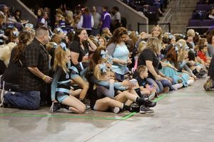 Young cheerleaders in matching teal and black uniforms sit on the arena floor beside parents and spectators at a cheer competition.