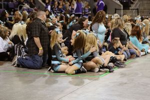 Young cheerleaders in teal and black uniforms sit on the floor with parents and a crowd of spectators watching a cheer competition.