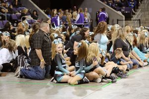 A crowd of spectators and cheerleaders in teal and black uniforms sit together on an arena floor during a cheerleading event.