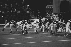 Black and white photo of cheerleaders performing a synchronized bending and kicking move on a competition floor with spectators behind.