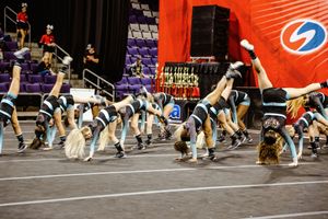 A cheer team performs a synchronized tumbling and leg-lift sequence on the arena floor while coaches watch from the sideline.
