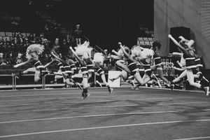 Black and white photo of a cheerleading squad leaping in unison with pom-poms during a performance in front of a seated crowd.