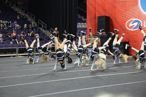 A cheer team in black and teal uniforms performs a synchronized tumbling routine across the mat in a large indoor arena.