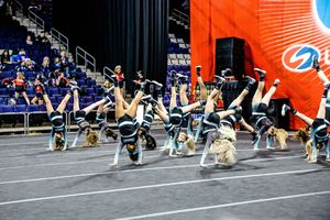 Cheerleaders perform a synchronized stunt on the mats, kicking their legs high in the air during a competition with bleachers behind them.