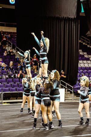 A cheerleading team in teal and black uniforms lifts a flyer into a stunt while teammates and spectators look on in a purple-seated arena.
