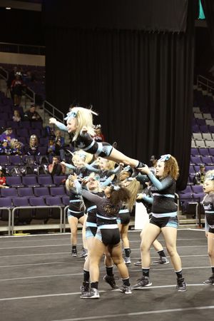Cheerleaders in black and teal uniforms lift a flyer high into the air during a stunt on the competition floor of an indoor arena.