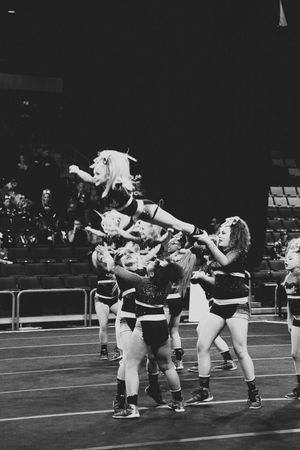 Black and white image of a cheerleader flying through the air in a basket toss as teammates reach up to catch her during a routine.