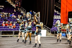 Cheer team in teal and black uniforms lifts flyers in a stunt sequence during a competition routine with an audience in the stands.