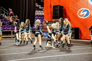 Cheerleaders in teal and black uniforms gather and perform choreography on the mat in front of an orange banner at a cheer competition.