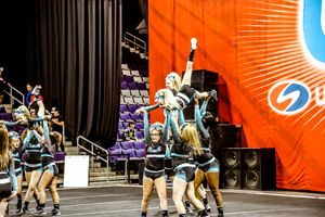 A flyer balances high in a one-footed extension stunt held by teammates in teal and black uniforms near an orange United Spirit banner.