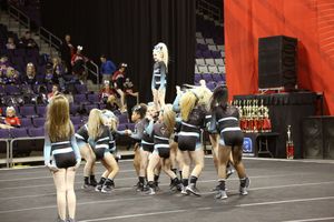 Cheerleading team in black and teal uniforms performs a stunt on the arena floor while spectators watch from the stands.