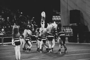 Black and white photo of cheerleaders lifting a flyer into a stunt while a teammate stands in the foreground at a cheer competition.