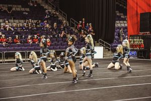A cheer team spreads across the competition mat in teal and black uniforms, performing choreography with spectators seated in the arena.