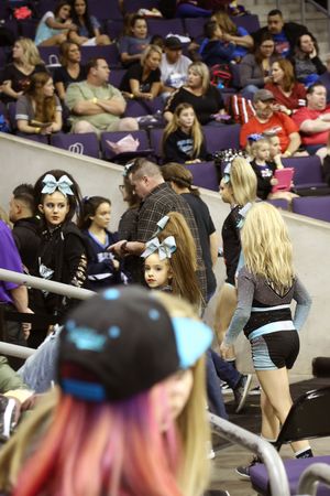 Young cheerleaders in black uniforms and bows walk along the arena bleachers as spectators watch the competition.